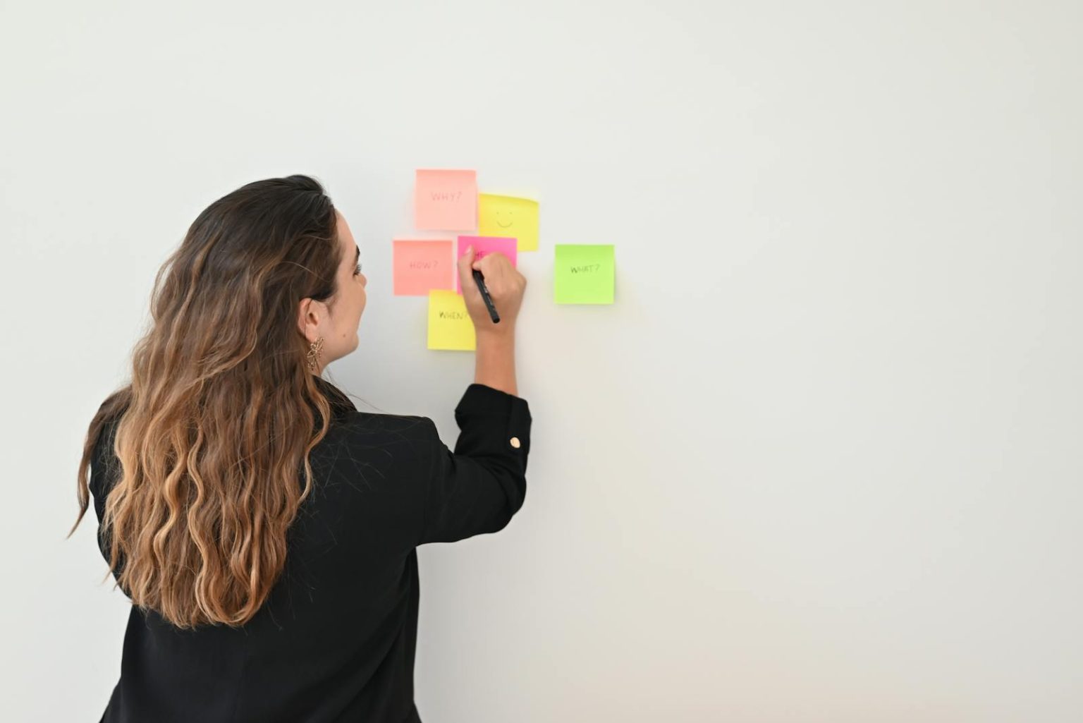 A businesswoman writes on colorful sticky notes on a white wall in an office setting.